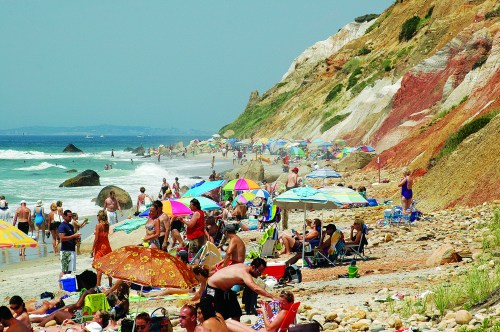 Crowded beach scene with colorful umbrellas, sunbathers, and rocky cliffs in the background.