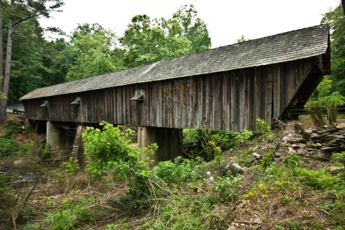 A rustic wooden covered bridge stands over a dry creek, surrounded by lush greenery and trees.