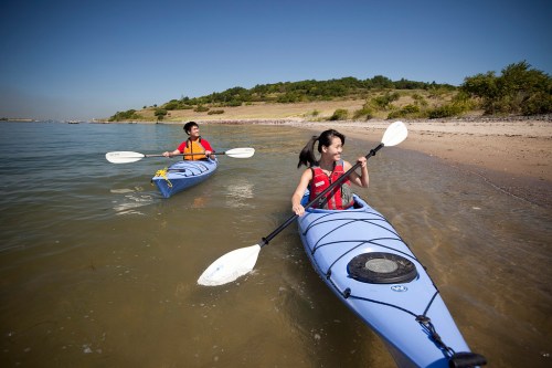 Two children paddle kayaks in calm water near a sandy shore on a sunny day. Green hills are visible in the background.