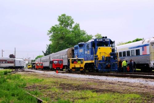 A blue and yellow train engine is parked next to several train cars in a yard, with workers and vehicles nearby.