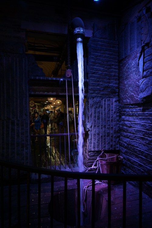 A water feature flows from a pipe into a wooden basin, illuminated by blue light in a rustic setting.