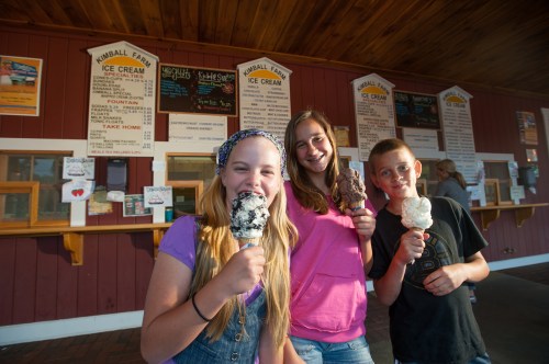 Three children smile while holding ice cream cones in front of an ice cream shop menu.