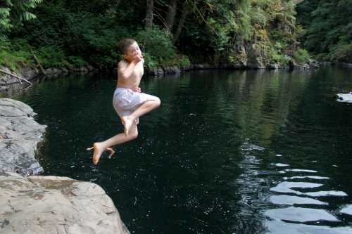 A boy jumps joyfully into a calm, green-tinted swimming hole surrounded by trees and rocky edges.