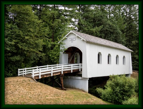 A white covered bridge stands over a grassy area, surrounded by lush green trees.