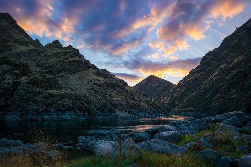 Sunset over a river surrounded by rugged mountains, with colorful clouds reflecting in the water.