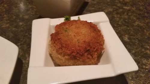 A golden-brown, crispy croquette served in a small white dish, garnished with a sprinkle of green herbs.