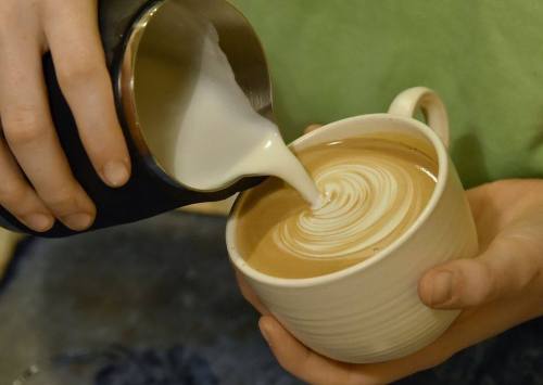 A person pours steamed milk into a coffee cup, creating a swirling latte art design.
