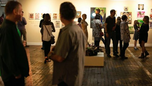 A crowded art gallery with people engaging and viewing colorful artworks on the walls.