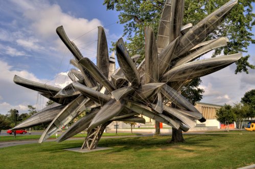 A large, abstract sculpture made of stacked canoes, resembling a tree, set in a grassy area with a cloudy sky.