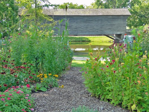 A scenic garden path lined with colorful flowers, leading to a pond and a wooden structure in the background.