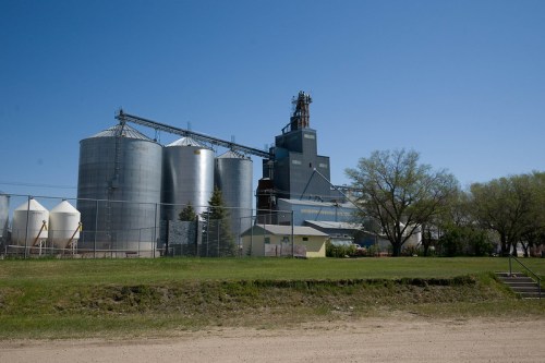 A grain elevator and silos against a clear blue sky, with green grass and trees in the foreground.