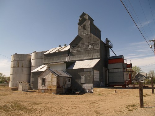A large, weathered grain elevator with silos and a small building in a rural setting under a clear blue sky.