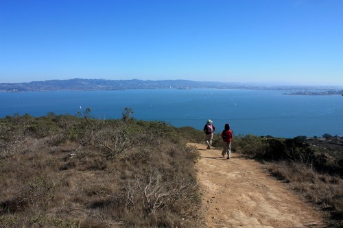 Two hikers walk along a dirt path overlooking a calm bay and distant hills under a clear blue sky.