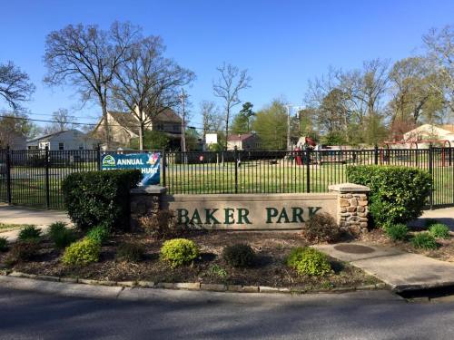 Sign for Baker Park with a playground in the background, surrounded by trees and houses under a clear blue sky.
