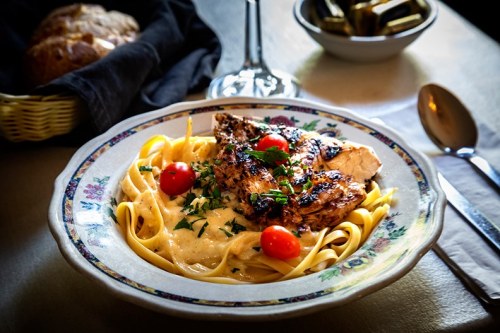 A plate of fettuccine topped with grilled chicken, cherry tomatoes, and herbs, with bread and a glass in the background.