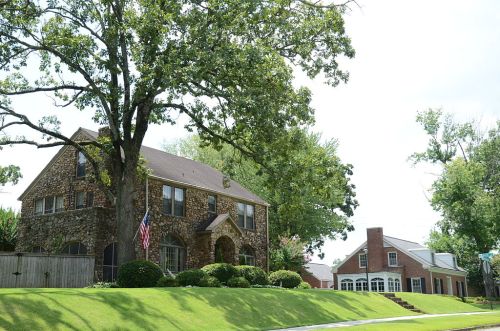 A stone house with an American flag, surrounded by greenery, next to a brick building on a sunny day.
