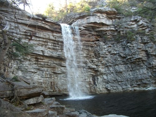 A serene waterfall cascading down rocky cliffs into a calm pool below, surrounded by trees and natural scenery.
