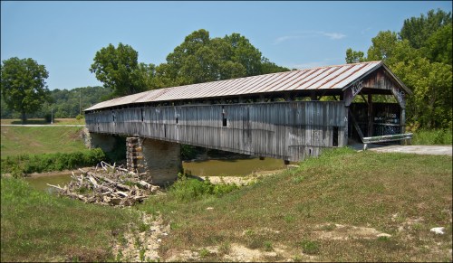 A weathered covered bridge spans a small creek, surrounded by lush greenery and a clear blue sky.