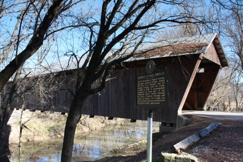 A wooden covered bridge with a sign, surrounded by trees and reflecting in the water below, under a clear blue sky.