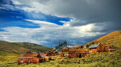 Abandoned wooden buildings in a ghost town, surrounded by green hills under a dramatic sky with clouds.