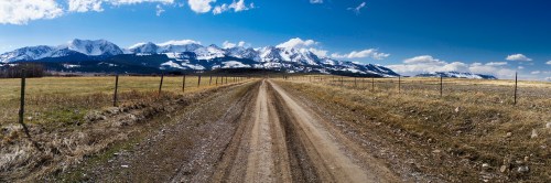 A dirt road stretches through a grassy field, leading to snow-capped mountains under a clear blue sky.