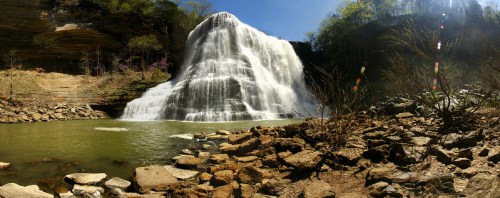A panoramic view of a cascading waterfall surrounded by rocky terrain and lush greenery under a clear blue sky.