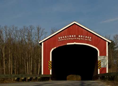 A red covered bridge with a sign reading "Buskirk's Bridge" surrounded by trees and a road sign indicating height and weight limits.