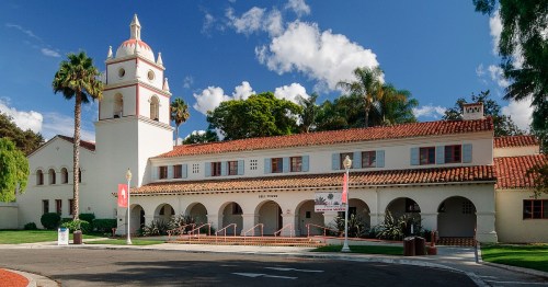 Historic building with a red-tiled roof, palm trees, and a clock tower under a blue sky with fluffy clouds.