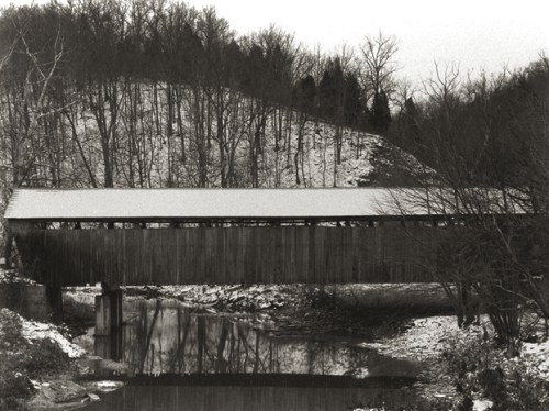 A black and white image of a covered bridge over a calm river, surrounded by snow-covered trees and a hillside.