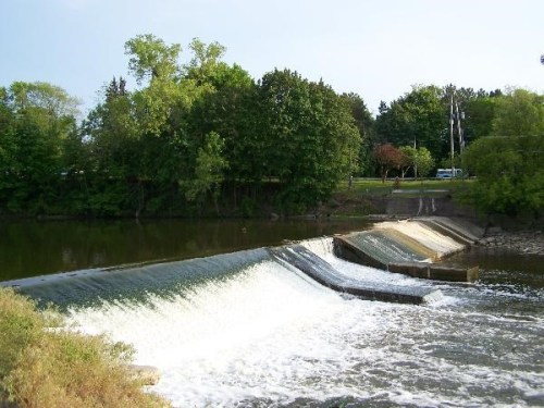 A serene river scene featuring a small dam with cascading water, surrounded by lush green trees and a clear sky.