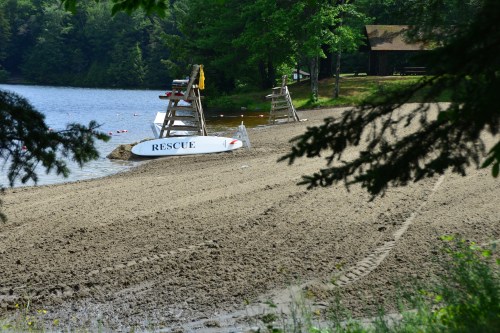 A sandy beach by a lake with a lifeguard stand labeled "RESCUE" and trees in the foreground.