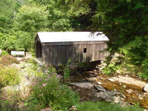 A rustic wooden covered bridge over a small stream, surrounded by lush greenery and colorful flowers.