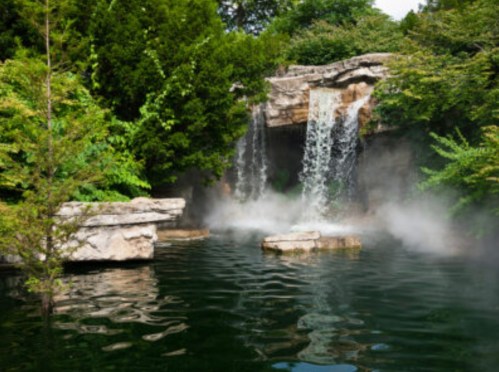 A serene waterfall cascades into a misty pond, surrounded by lush greenery and rocky formations.