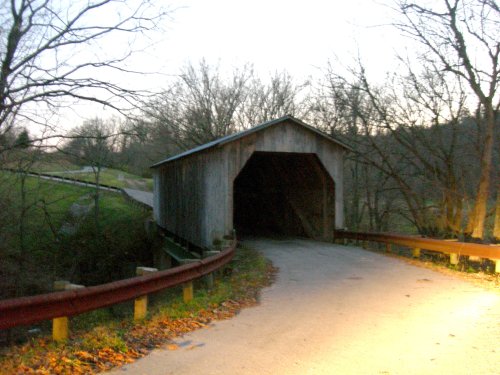 A rustic covered bridge at dusk, surrounded by bare trees and a winding road.