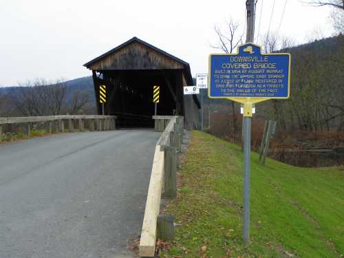 A covered bridge with a historical sign, surrounded by trees and a rural landscape on a cloudy day.