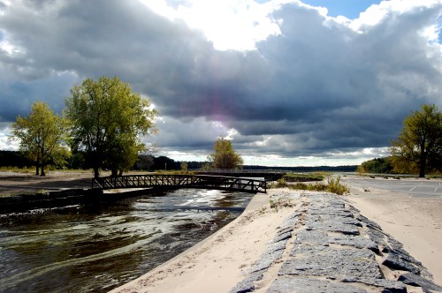 A wooden bridge over a river, surrounded by trees and a sandy bank, under a cloudy sky.
