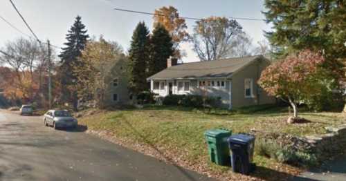 A suburban house with a front yard, surrounded by trees and parked cars on a quiet street in autumn.