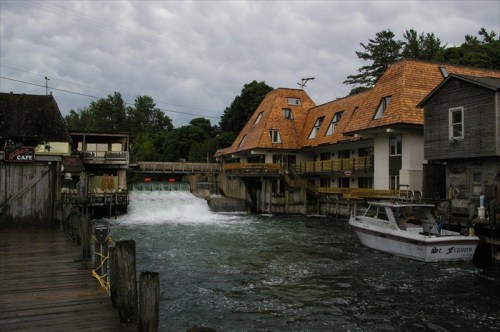 A scenic view of a waterway with a small boat, a dam, and buildings featuring unique sloped roofs under a cloudy sky.