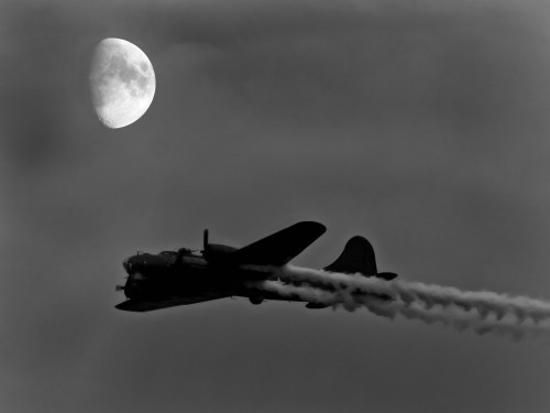 A vintage airplane flies against a large moon, leaving a trail of vapor in a dramatic black and white sky.