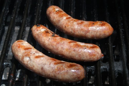 Three grilled sausages sizzling on a barbecue grill, showcasing a golden-brown exterior with grill marks.