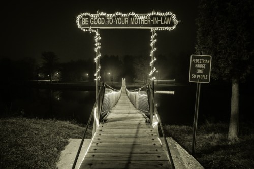 A lit bridge at night with a sign reading "Be Good to Your Mother-in-Law" and a pedestrian limit sign nearby.