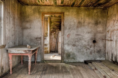 An empty, weathered room with a wooden table, peeling walls, and a doorway leading to another space.