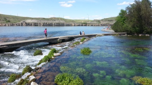 A serene river scene with people walking on a path, lush greenery, and clear water reflecting the sky.