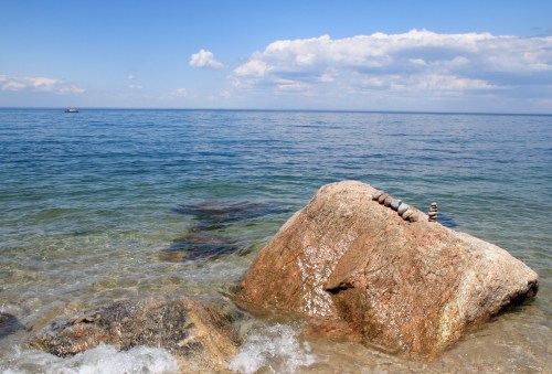A large rock in shallow water with a clear blue sky and calm sea in the background.