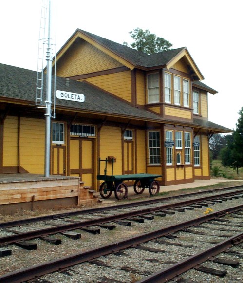 Historic Goleta train station with yellow exterior, large windows, and a small cart on the platform beside the tracks.
