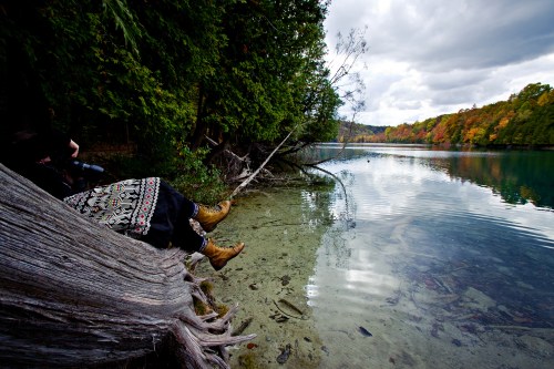 A person relaxes by a serene lake, surrounded by trees and autumn foliage, with their feet resting on a log.