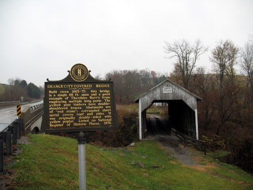 A historic covered bridge with a sign detailing its history, surrounded by trees and a rural landscape.