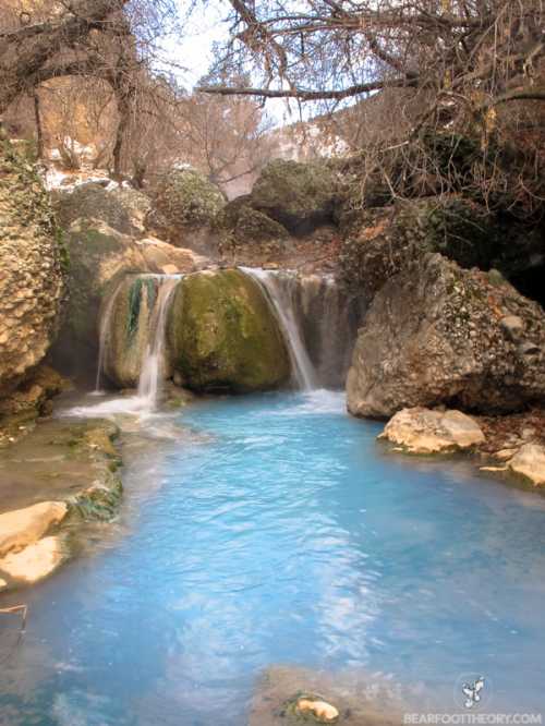 A serene scene of a blue river flowing over rocks, surrounded by trees and boulders in a tranquil natural setting.