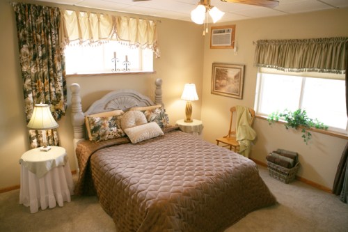 Cozy bedroom with a quilted bedspread, lamps, and warm decor, featuring a window with curtains and a chair.