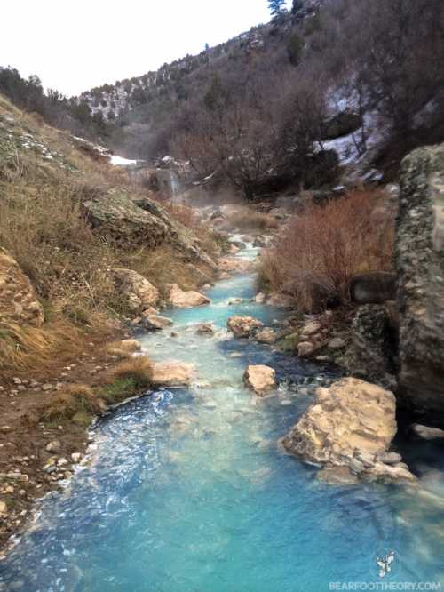 A serene, turquoise stream flows through rocky terrain, surrounded by bare trees and a misty atmosphere.